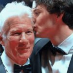 Dos hombres vestidos de gala en una alfombra roja; uno besa la mejilla del otro en un gesto afectuoso.