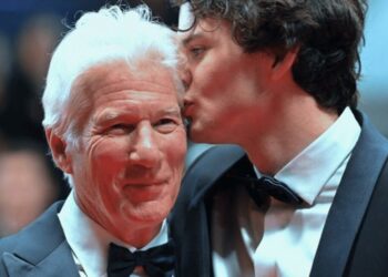 Dos hombres vestidos de gala en una alfombra roja; uno besa la mejilla del otro en un gesto afectuoso.