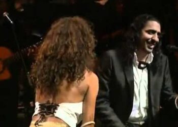 Dos artistas en el escenario, mujer de espaldas con vestido blanco y hombre sonriente con traje negro