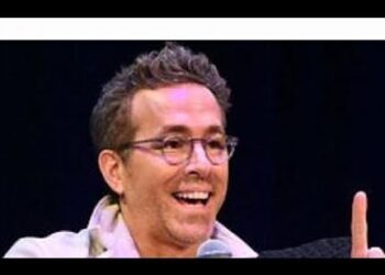 Hombre adulto con gafas sonriendo y hablando en un escenario con micrófono, gesto animado.