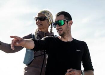 Dos personas con gafas deportivas y ropa de montaña señalando hacia el horizonte en un paisaje luminoso