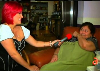 Entrevistadora con cabello rojo sostiene un micrófono frente a una mujer sentada en un sofá en un ambiente doméstico — ¿Quién es la esposa de Pedro Sevcec? Descubre su historia y más