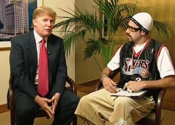 Dos hombres sentados frente a frente en una oficina, uno con traje formal y corbata y otro con vestimenta excéntrica y gorra, en una escena que sugiere una entrevista: ¿De dónde es Borat? Descubre la verdad sobre su origen kazajo