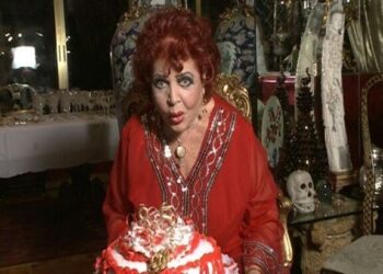 Mujer mayor de maquillaje marcado y vestido rojo sentada frente a un pastel y objetos decorativos en una sala opulenta, La sorprendente vida de los hijos de Sara Montiel: Thais y Zeus