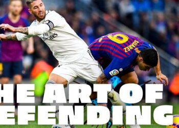 Dos futbolistas en intensa disputa por el balón en un estadio, entrada defensiva con texto superpuesto