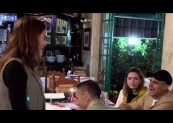 La familia de Yuya: secretos, momentos y la vida tras la fama en una cena íntima, con una mujer de pie conversando y varios familiares sentados alrededor de una mesa en un restaurante acogedor.