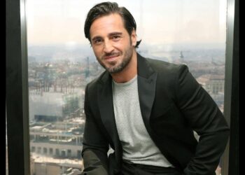 Hombre adulto con chaqueta negra y barba sentado junto a un gran ventanal con vistas urbanas, imagen sin identificación