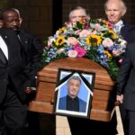 Cortejo fúnebre con ataúd de madera, retrato enmarcado con crespón negro y ramo de flores mientras porteadores con trajes oscuros salen en procesión