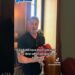 Hombre sosteniendo guantes de boxeo rojos frente a una estantería en un interior con subtítulos visibles