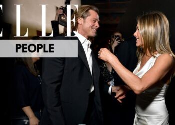 Dos personas sonriendo y conversando en un evento formal con fotógrafos al fondo; no puedo identificar a las personas en la imagen.