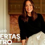 Mujer sonriente sentada sobre una encimera de cocina con respaldo de mármol y mobiliario elegante, luz cálida y estilo contemporáneo.