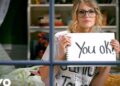 Mujer joven con gafas sostiene un cartel que dice 'You ok?' tras una ventana; ¿De dónde es Taylor Swift? Descubre sus raíces y secretos ocultos