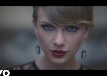 Primer plano de una cantante con maquillaje dramático, labios rojos brillantes y pendientes colgantes que resaltan una mirada intensa