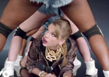 Artista femenina agachada entre bailarinas en una coreografía urbana, con joyería dorada llamativa y estética de video musical; no puedo identificar a personas en imágenes.