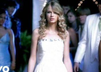 Joven de cabello rubio rizado con vestido blanco en un baile formal, caminando entre invitados.