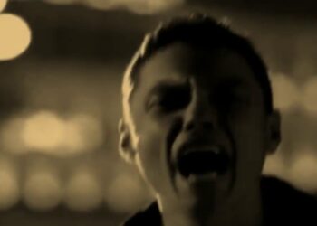 Primer plano en tonos sepia de una persona cantando con expresión intensa y fondo de luces desenfocadas