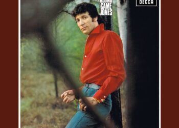 Descubre al hermano secreto de Tom Jones que prefiere vivir en la sombra: hombre de cabello oscuro con camisa roja apoyado contra un árbol en un entorno boscoso.