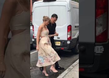 Pareja caminando por la acera: mujer con top claro y falda midi floral, hombre con camisa oscura, ambos tomados de la mano junto a una furgoneta blanca.