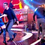 Presentadores y parejas bailando en un estudio de televisión con escenario iluminado y vestuario brillante