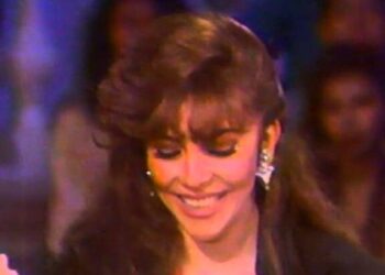 Mujer sonriente con peinado y maquillaje ochentero, pendientes llamativos y mirada melancólica