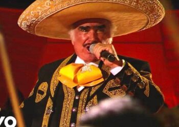 Cantante de mariachi en traje charro negro con bordados dorados, gran sombrero mexicano y moño amarillo sosteniendo un micrófono en el escenario