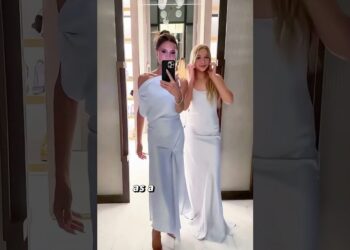 Dos mujeres con vestidos largos blancos posando frente a un espejo en un vestidor, transmitiendo elegancia y estilo.