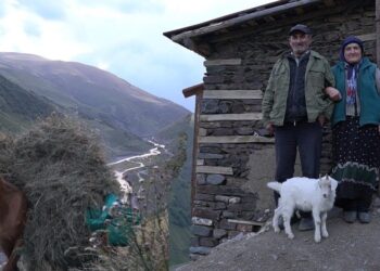 Pareja de granjeros junto a su cabra frente a una casa de piedra en un valle montañoso con un caballo cargado de heno