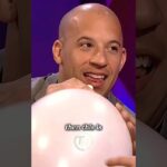 Hombre calvo sonriente sosteniendo un globo rosa en un plató con fondo morado.