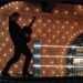 Silueta de guitarrista sobre un escenario iluminado con pared de luces y estantería de botellas — La sorprendente fortuna de Antonio Banderas: ¿cuánto tiene realmente?