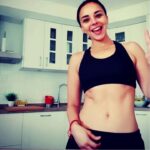 Mujer sonriente en ropa deportiva posando en una cocina moderna con encimera de madera y azulejos blancos.
