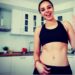 Mujer sonriente en ropa deportiva posando en una cocina moderna con encimera de madera y azulejos blancos.