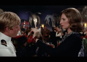 Mujer sosteniendo una copa y hablando con un hombre de uniforme en una elegante sala de fiesta; Descubre el thriller de 8 episodios en Prime Video que todos están viendo y que está basado en un bestseller ¡No podrás parar de verlo!