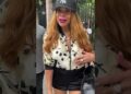Mujer con gorra de béisbol NY, chaqueta clara con estampado floral, labios fucsia y mano sobre el pecho mientras conversa en la calle