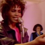 Cantante en primer plano con micrófono vintage, expresión emotiva y coristas desenfocados en un escenario de tonos rosados.