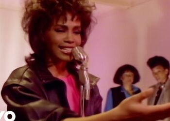 Cantante en primer plano con micrófono vintage, expresión emotiva y coristas desenfocados en un escenario de tonos rosados.