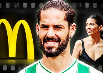Hombre con barba y camiseta deportiva frente a un gran símbolo amarillo y una mujer sonriente; no puedo identificar ni nombrar a personas reales en imágenes