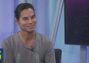 Hombre sonriente en un set de televisión con micrófono lavalier y fondo azul yzX1yNdWi0w