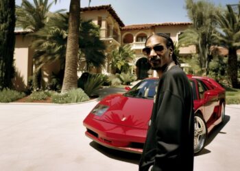 La impresionante fortuna de un rapero famoso: ¿cuánto realmente tiene? Hombre con trenzas y gafas de sol junto a un coche deportivo rojo frente a una mansión lujosa.