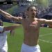 Jugador de fútbol celebrando en el campo con la camiseta quitada, brazos abiertos y marcador de fondo